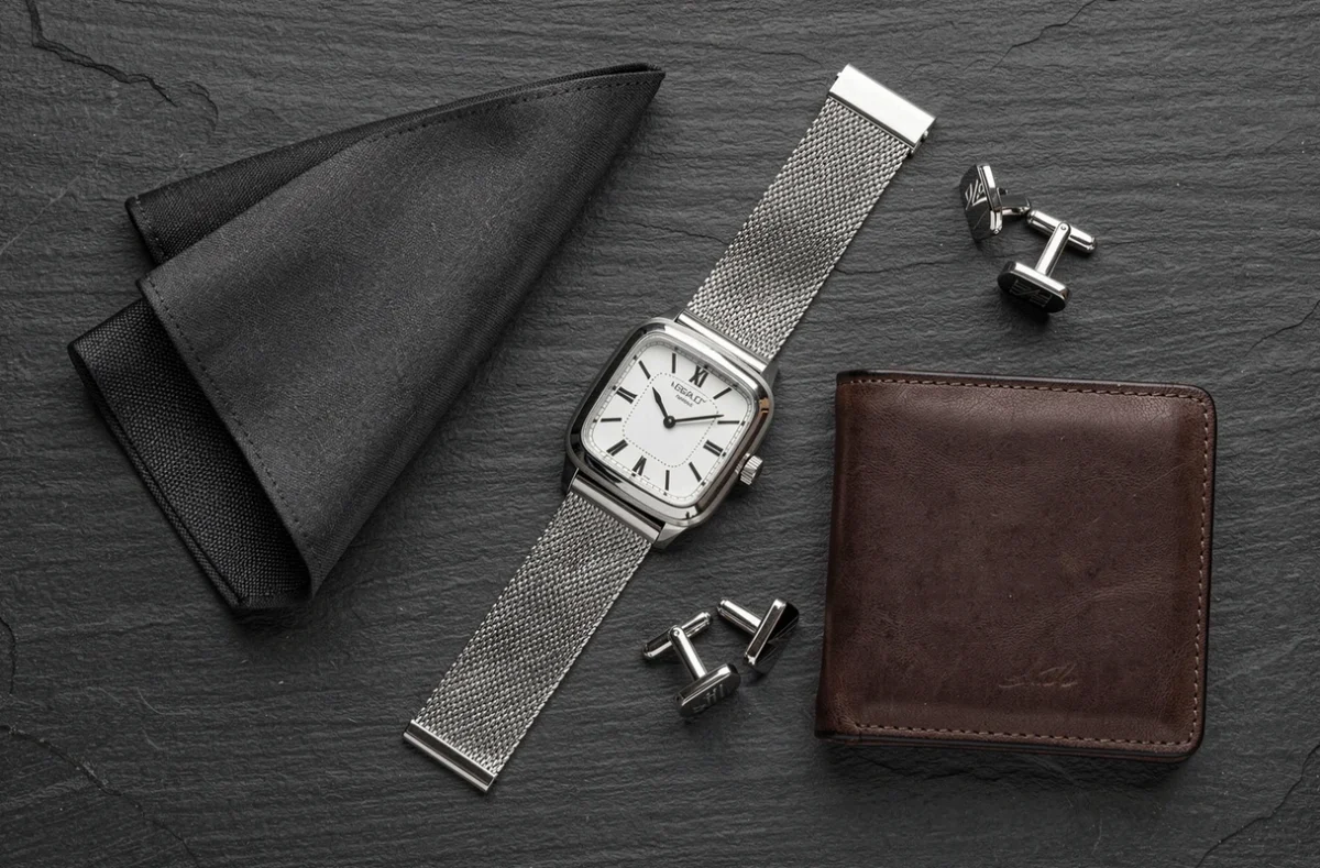 Flat-lay élégant d'une montre carrée en acier avec bracelet maille sur ardoise grise, accompagnée d'accessoires de mode masculine
